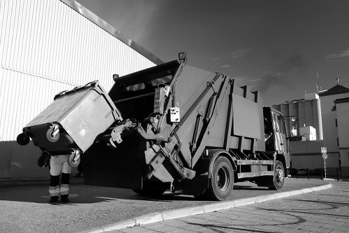 A large industrial waste collection vehicle with a hydraulic lifting arm is engaged in lifting a wheeled rubbish bin positioned at the rear of the truck. The bin appears to be made of plastic with a textured surface and is mounted on wheels, with its lid closed. The vehicle is parked on a paved area adjacent to a tall, corrugated metal building, possibly a warehouse or commercial premises in Westminster. The truck has a predominantly dark body with visible mechanical components on the arm, and the environment suggests an outdoor commercial setting with a partly cloudy sky overhead. The image captures the process of rubbish collection as part of waste management services offered by Commercial Waste Westminster, emphasizing the handling of waste bins in a business or industrial context within the Westminster area, inferred from the webpage details.