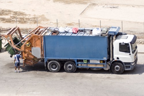 A large waste collection truck with a white cab and blue rear container is parked on a paved road in an open area, possibly near a construction site or industrial zone. The truck is actively engaged in rubbish removal, with some waste bags and miscellaneous debris loaded into the top of the container. Two workers, one wearing a blue shirt and the other a white shirt, are positioned at the front of the truck near the hopper, likely assisting with loading or maintenance. The waste material appears to include plastic bags, cardboard, and other miscellaneous refuse, suggesting a general rubbish collection operation. The environment around the truck includes a dirt or gravel surface, with a chain-link fence and an area of exposed ground visible in the background. This scene is typical of commercial waste collection services offered by companies like Commercial Waste Westminster, operating in the Westminster area, and illustrates professional rubbish removal in an urban or semi-urban setting, emphasizing the practical handling of business waste in the local vicinity.