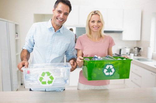 A man and a woman stand in a bright, modern kitchen holding clear and green recycling bins with white recycling symbols printed on the sides. The man, dressed in a light blue shirt, holds a transparent bin containing various recyclable plastics and paper materials, while the woman, wearing a pink top, presents a green bin filled with similar recyclable waste. Behind them, the kitchen features white cabinets, a stainless steel stove, and a few kitchen utensils visible on the counter. The scene appears well-lit with natural light, emphasizing the cleanliness and organization typical of a domestic recycling setup. This image relates to rubbish management and recycling services, aligning with the services offered by Commercial Waste Westminster, especially in the context of promoting proper waste segregation within residential environments in Westminster, SW1 postcode area.