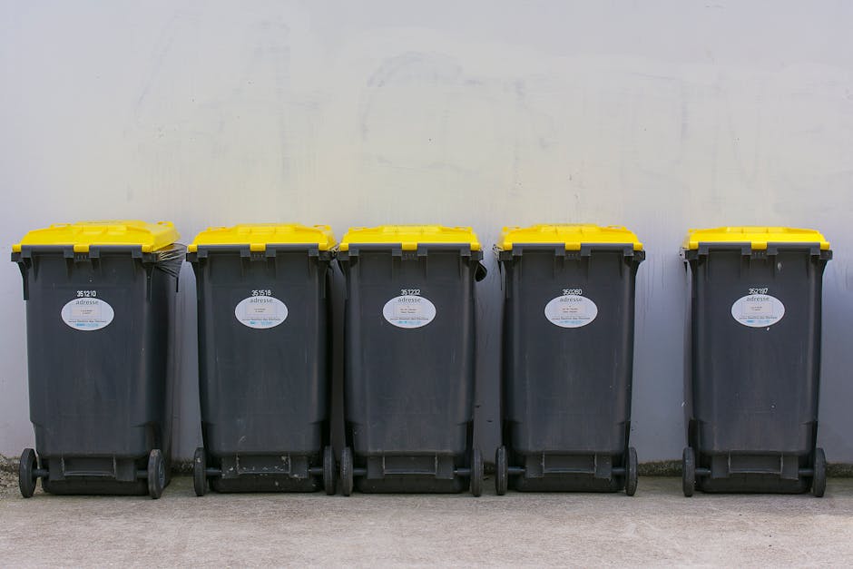 Five large wheeled rubbish bins made of dark grey durable plastic with textured surfaces, positioned in a row against a plain, light-colored wall. Each bin has a bright yellow lid, some with slightly