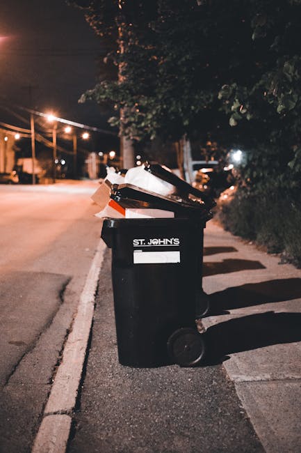 Five large wheeled rubbish bins made of dark grey durable plastic with textured surfaces, positioned in a row against a plain, light-colored wall. Each bin has a bright yellow lid, some with slightly